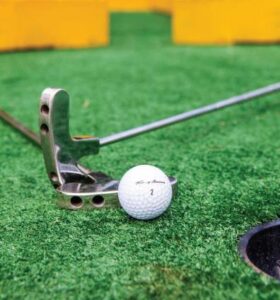 Una pelota de golf en Pasto Sintético Golfito cerca de un hoyo con un putter colocado detrás.