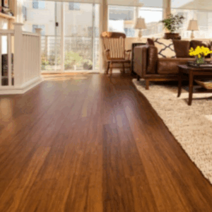 Salón soleado con suelo de Piso en Bambú Macizo Caramelo, alfombra beige, sofá marrón, mecedora, mesa de centro y grandes ventanales con vistas al exterior.