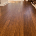 Salón soleado con suelo de Piso en Bambú Macizo Caramelo, alfombra beige, sofá marrón, mecedora, mesa de centro y grandes ventanales con vistas al exterior.