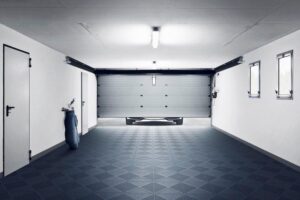 Una persona se encuentra en un garaje limpio y vacío con una puerta enrollable cerrada, rodeado de paredes y puertas blancas, y un Piso de Polipropileno Propósito debajo de unas pequeñas ventanas.