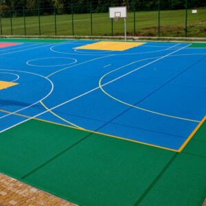 Una cancha de baloncesto al aire libre vacía y colorida del Piso de Polipropileno Multi Deportivo con múltiples líneas y zonas marcadas, rodeada por una cerca y un área de césped.