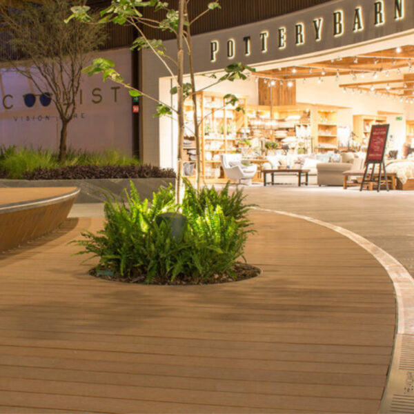 Una moderna zona comercial al aire libre con suelos Piso Deck WPC Redondo, jardineras y entradas a tiendas al atardecer.