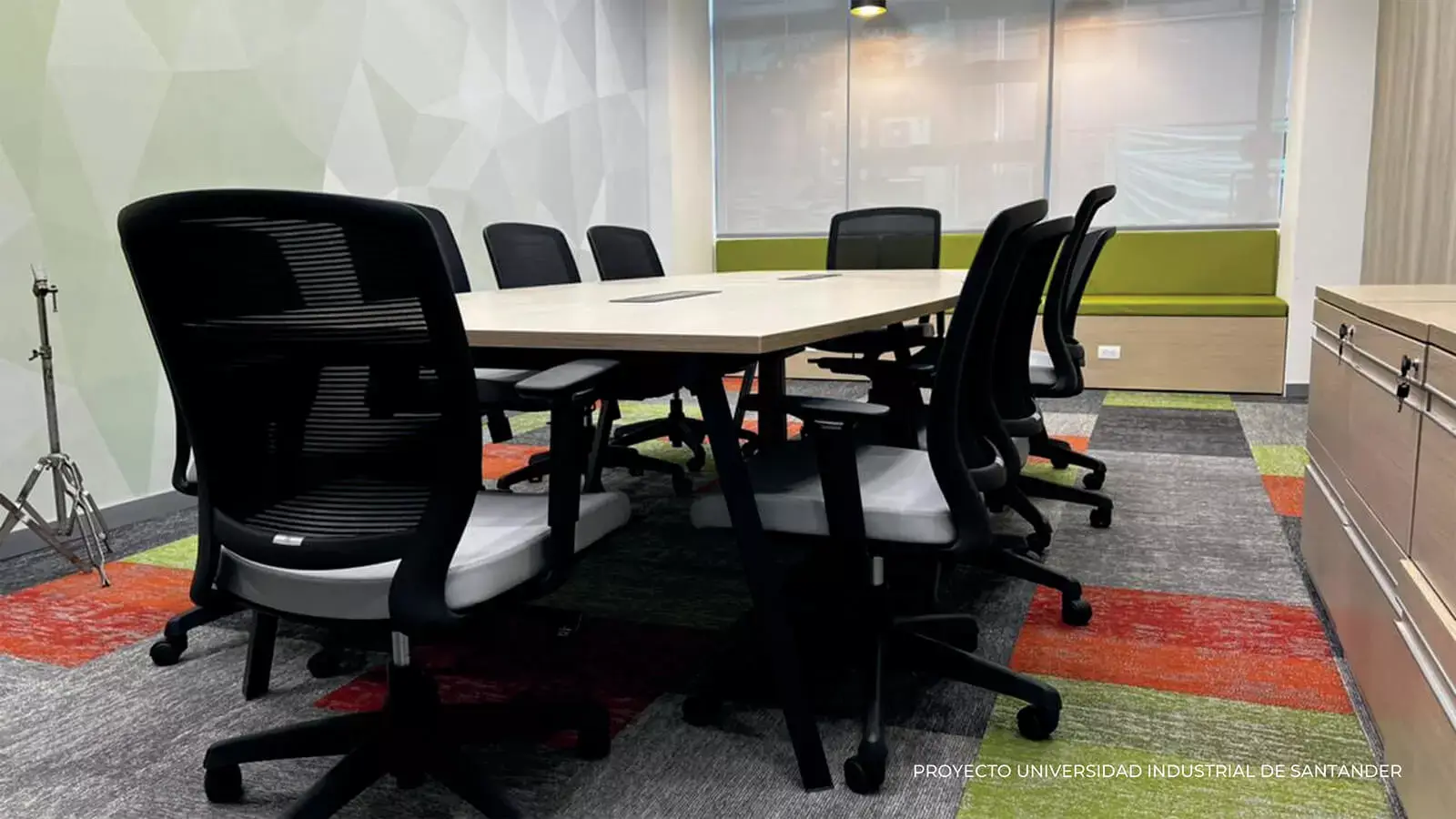 Moderna sala de conferencias con una gran mesa rectangular, rodeada de sillas de oficina negras, una alfombra colorida y un armario verde.