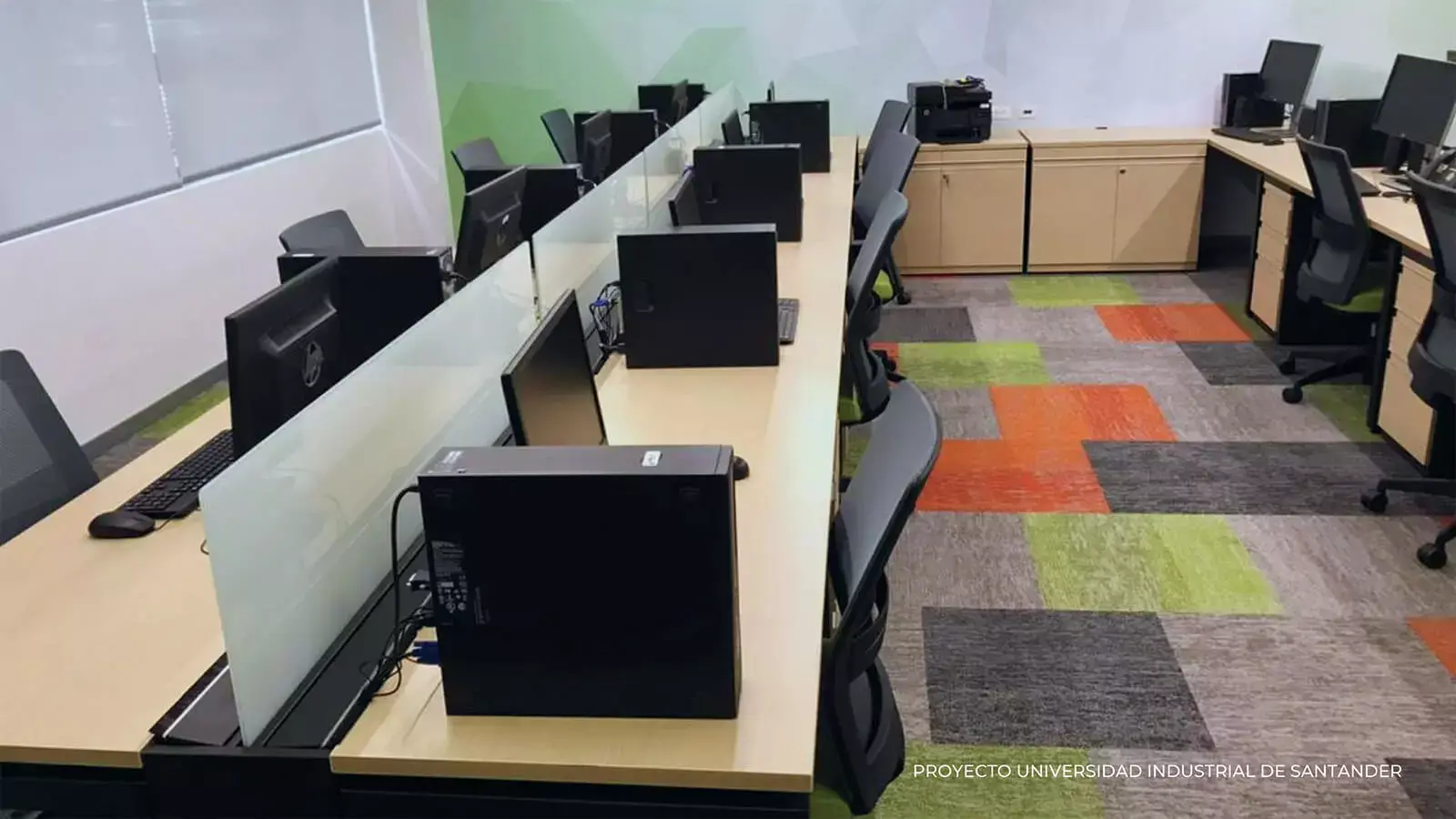 Oficina moderna con múltiples estaciones de trabajo, cada una con una computadora de escritorio y divididas por mamparas verdes, frente a un piso alfombrado de colores.