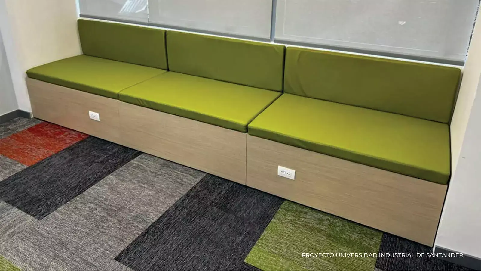Moderna sala de espera de oficina con un largo banco verde y gris con almacenamiento incorporado, frente a una vibrante alfombra multicolor.