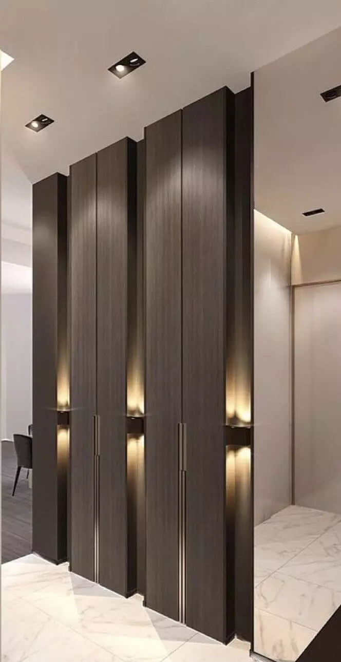 Pasillo moderno con paneles altos de madera oscura e iluminación incorporada, suelo de baldosas de mármol blanco y un atisbo de una habitación con muebles oscuros al fondo.