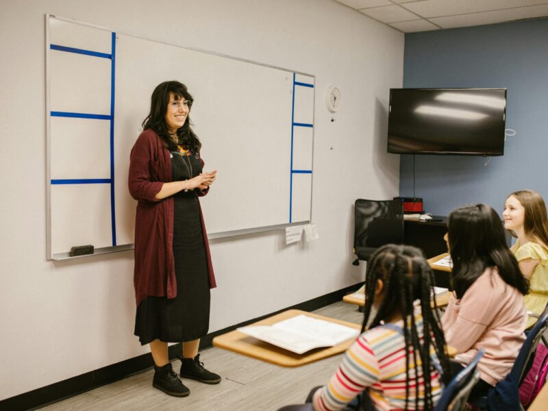Una mujer se sitúa al frente de un aula y habla a cuatro alumnos sentados con cuadernos abiertos en sus pupitres.