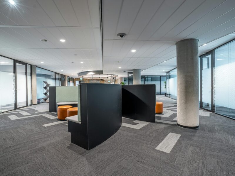 Moderno espacio de oficinas con salas de reuniones con paredes de cristal, moqueta gris, lámparas de techo y una zona central de asientos con tabiques negros y taburetes marrones.