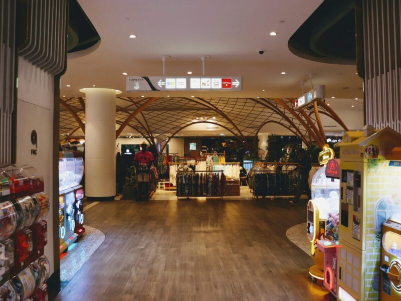 Pasillo de un centro comercial con máquinas expendedoras a ambos lados, suelo de madera, señalización aérea y estantes de ropa en el fondo central.