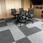 Una pequeña sala de conferencias con una mesa negra, cuatro sillas negras de rejilla, paredes con paneles de madera y baldosas de moqueta con motivos grises queda realzada por la entrada modular Alfombra, que la hace ideal para reuniones de equipo.
