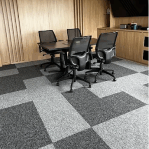 Una pequeña sala de conferencias con una mesa negra, cuatro sillas negras de rejilla, paredes con paneles de madera y baldosas de moqueta con motivos grises queda realzada por la entrada modular Alfombra, que la hace ideal para reuniones de equipo.